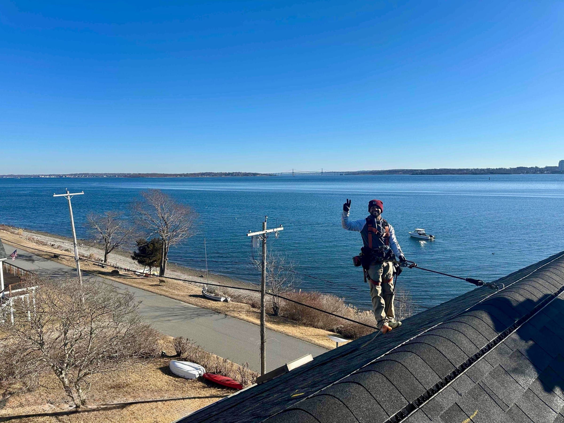 A crew member in a harness stands on the ridge of a black architectural-shingle roof above a wide bay with a long bridge on the horizon.