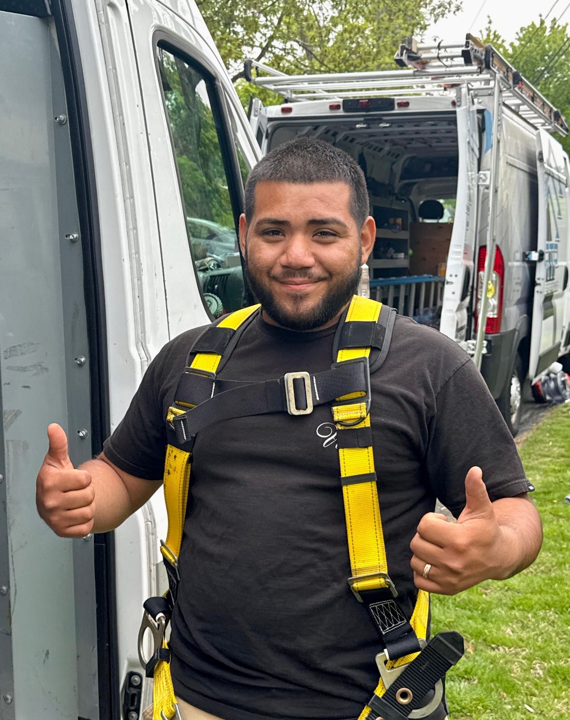 A crew member in a harness gives a thumbs-up beside two white work vans on a grassy lot.