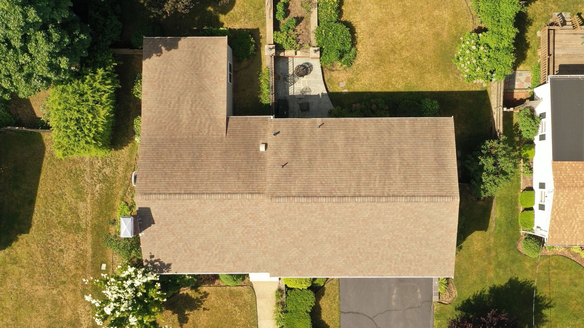 Overhead drone view of a worn tan shingle roof beside a patio and lawn.