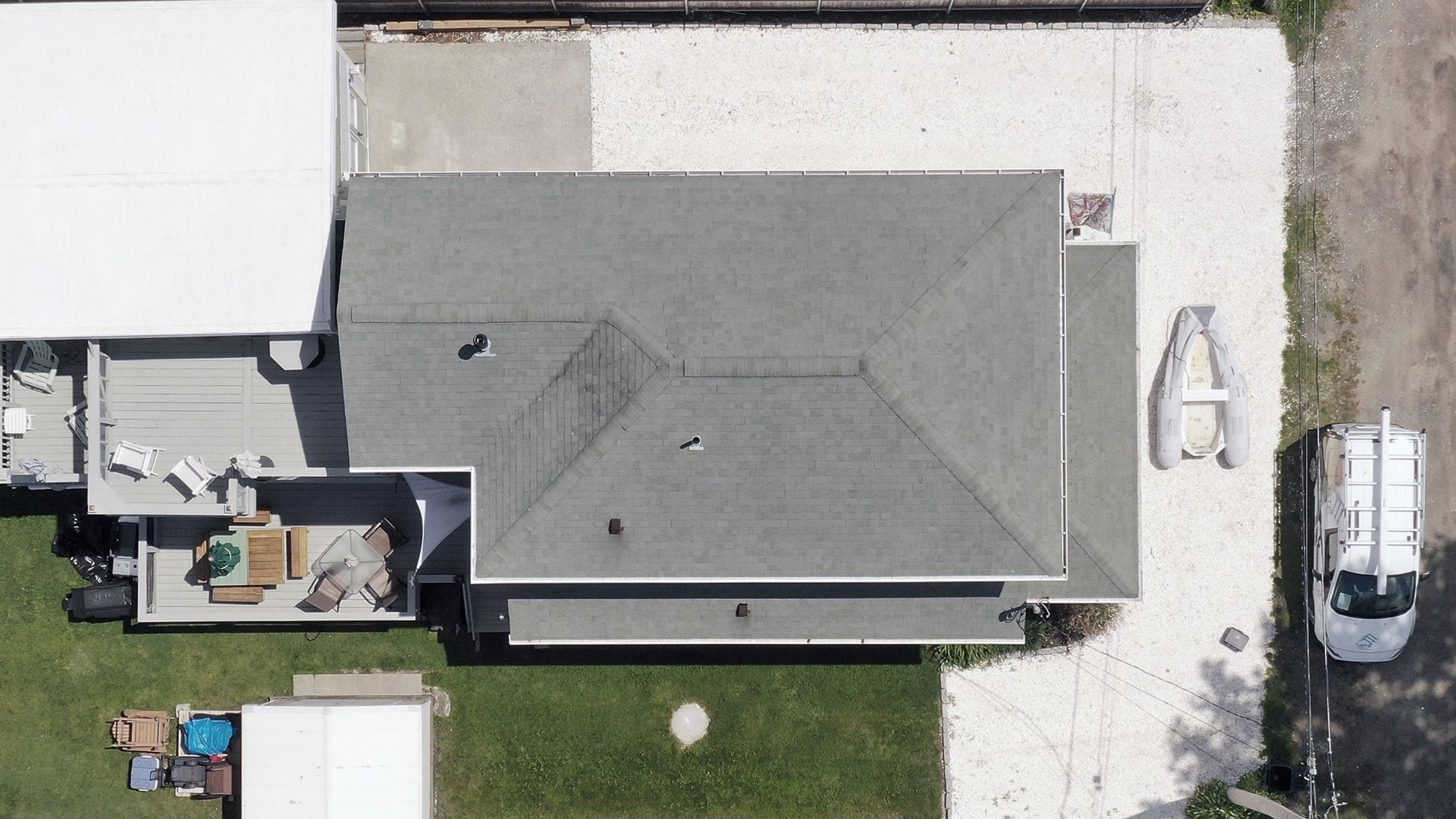 Overhead view of a weathered grey-green hip roof beside a gravel lot and a work van.