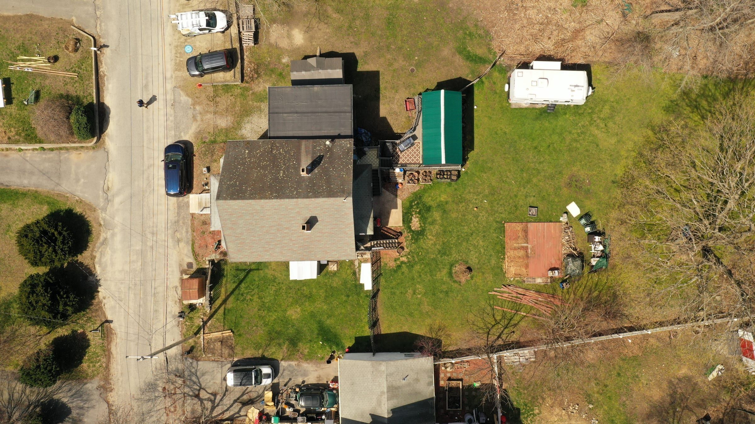 Wide drone view of a moss-spotted dark roof with two chimneys on a residential street.
