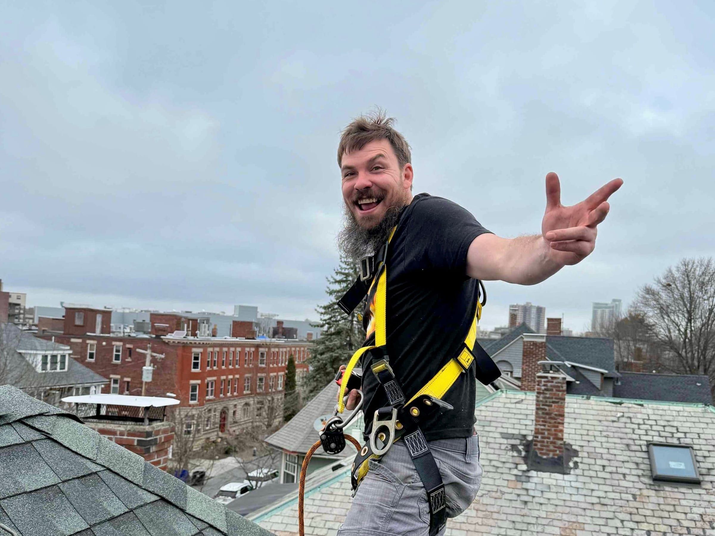 A roofer in a harness stands at the ridge of a grey shingle roof above a dense neighborhood of brick and clapboard rooftops.