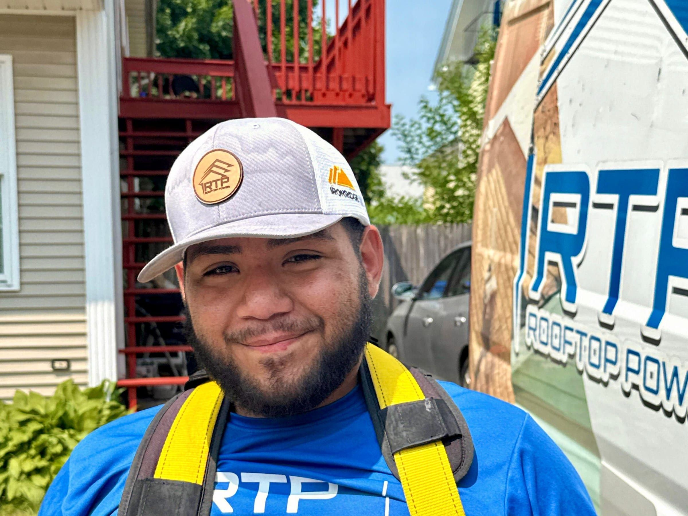 A roofer in a blue work shirt and yellow safety harness smiles beside a work van.