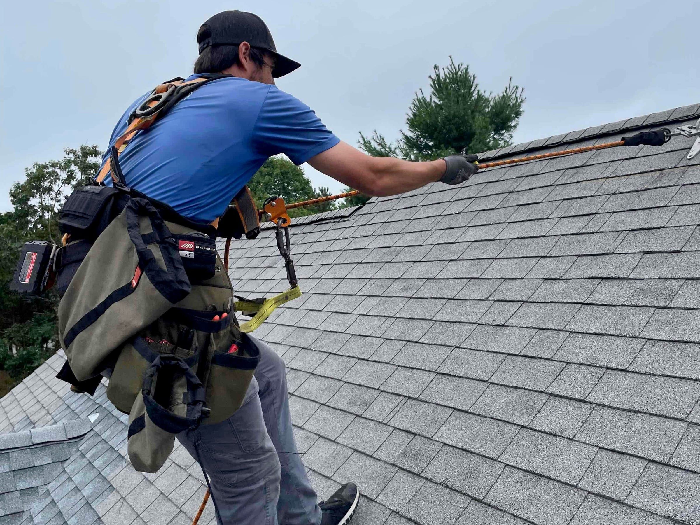 A roofer in a harness feeds a safety rope up a grey asphalt shingle roof toward a ridge anchor under an overcast sky.