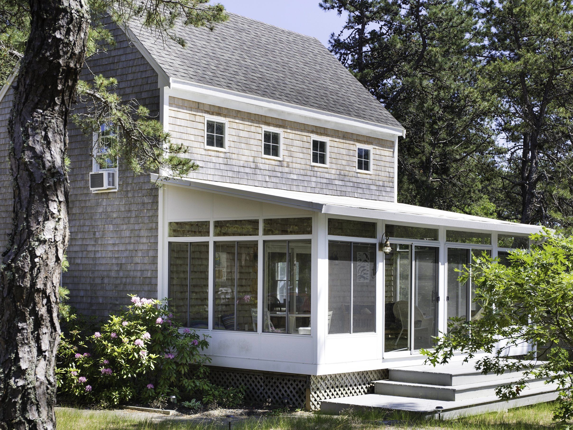 A weathered cedar-shingle house with a screened porch under pine trees.