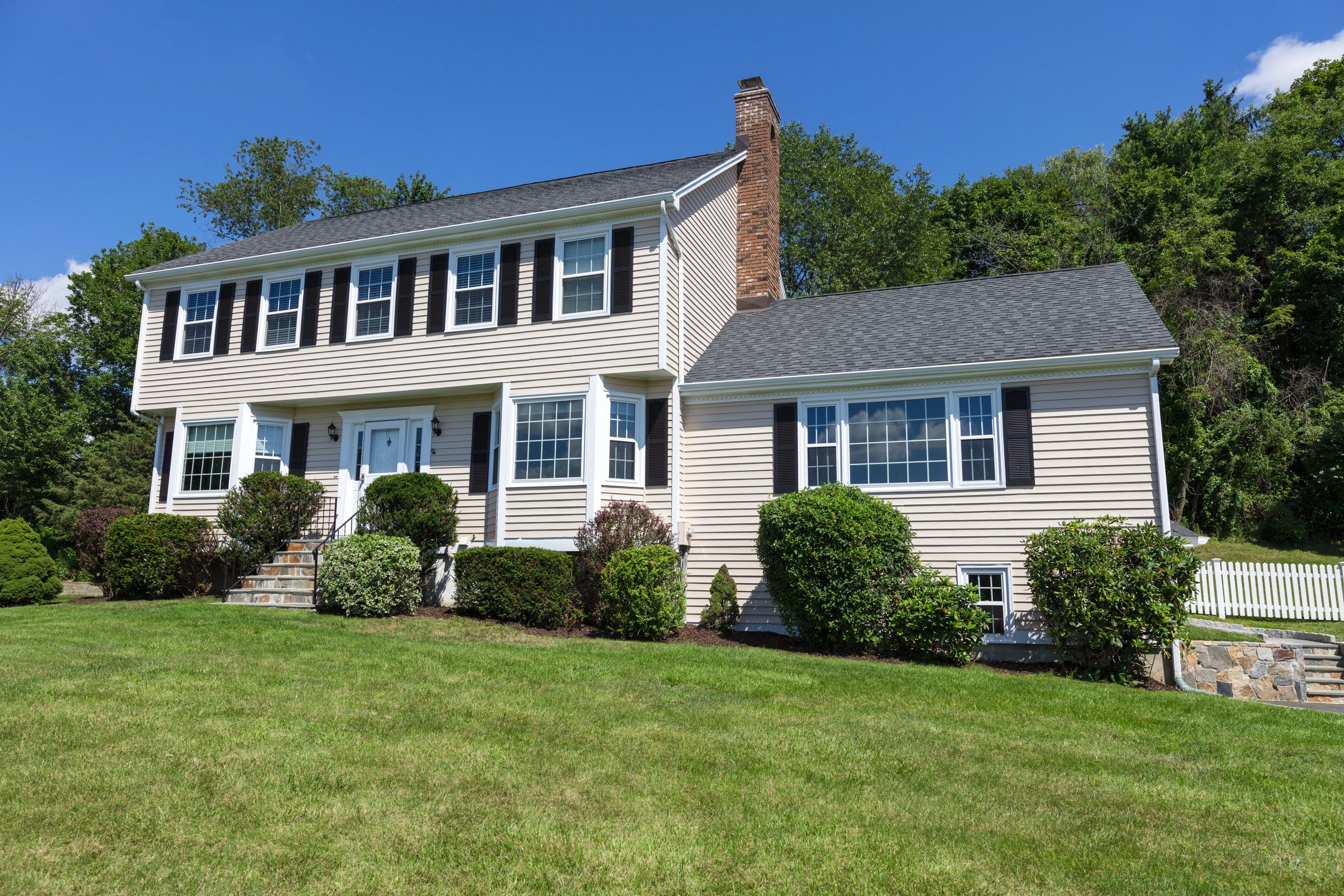A beige colonial with black shutters, a brick chimney and a grey shingle roof on a wide lawn.