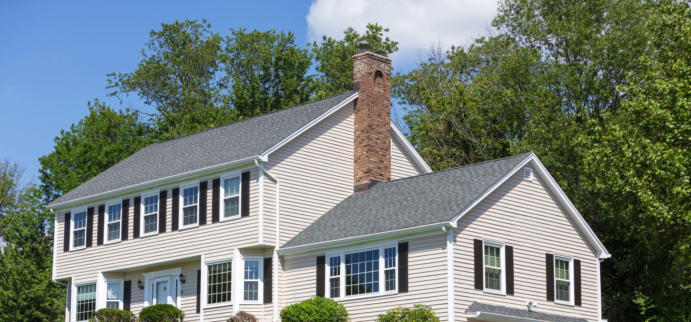 A grey-sided colonial with a brick chimney and a two-car garage on a hillside lawn.