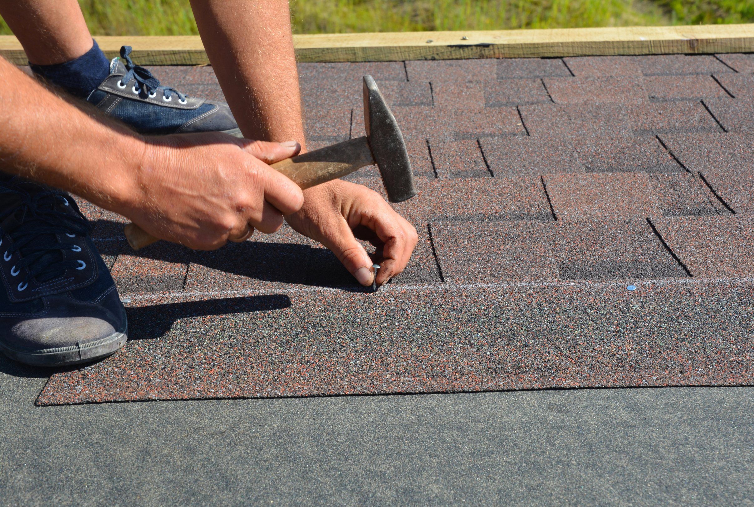 Hands hammering a nail into a new course of brown shingles.