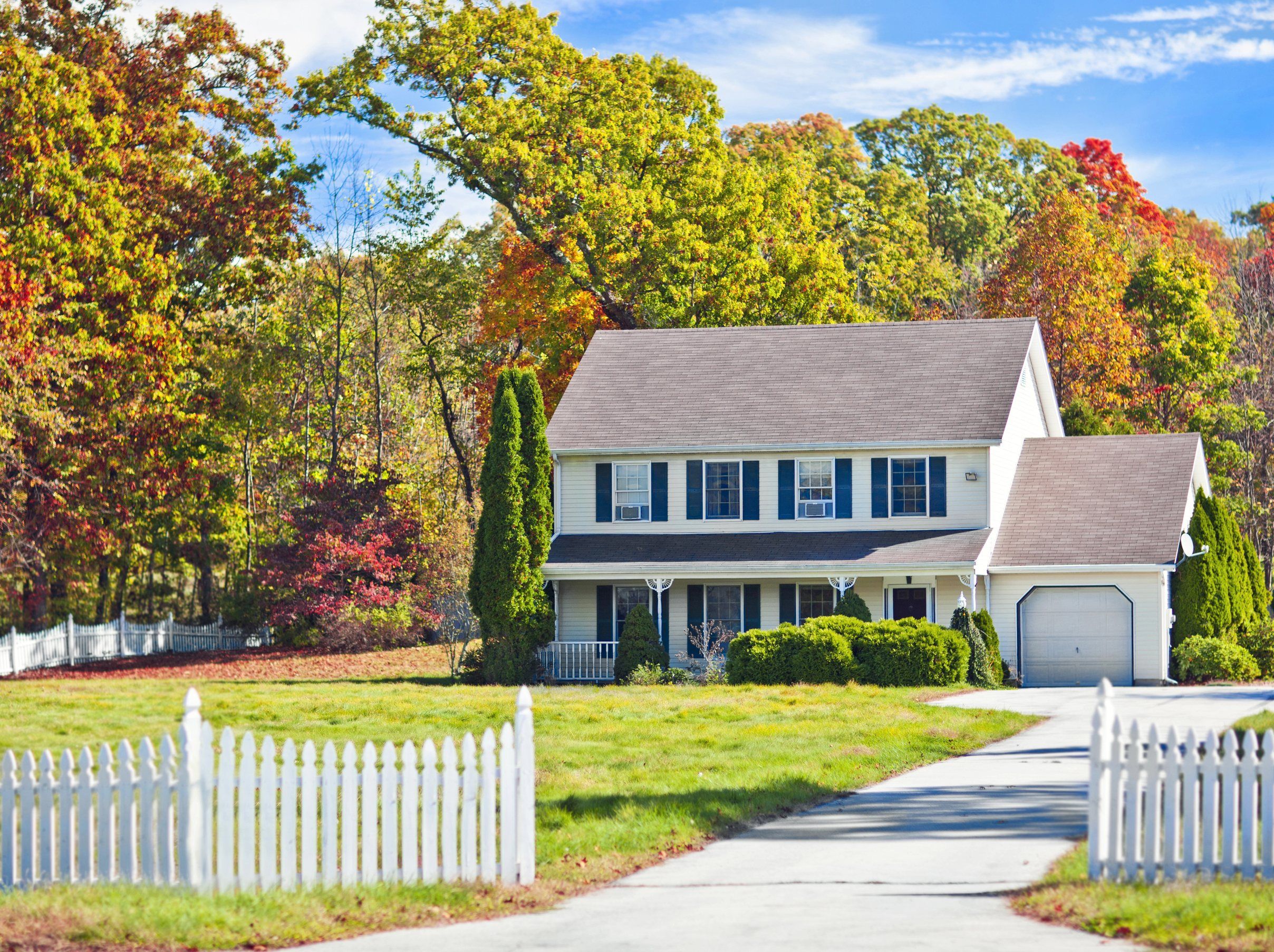 A white colonial with blue shutters and an attached garage under autumn trees behind a picket fence.