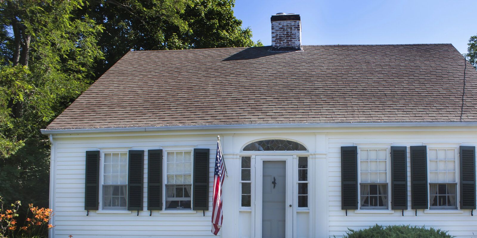 A white clapboard cape house with black shutters and an older, faded shingle roof with a brick chimney.