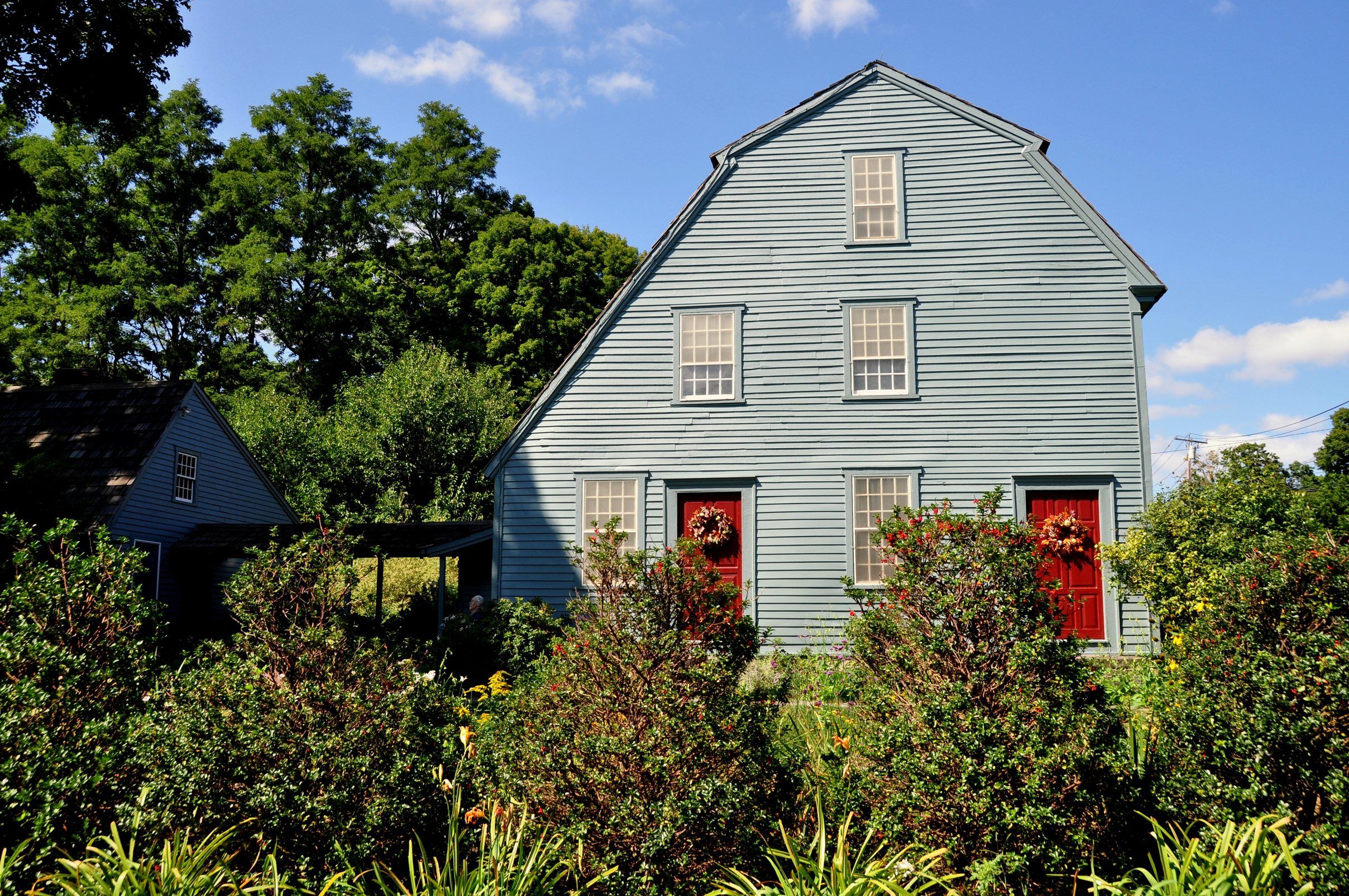 A blue-grey clapboard gambrel house with two red doors behind a summer garden.