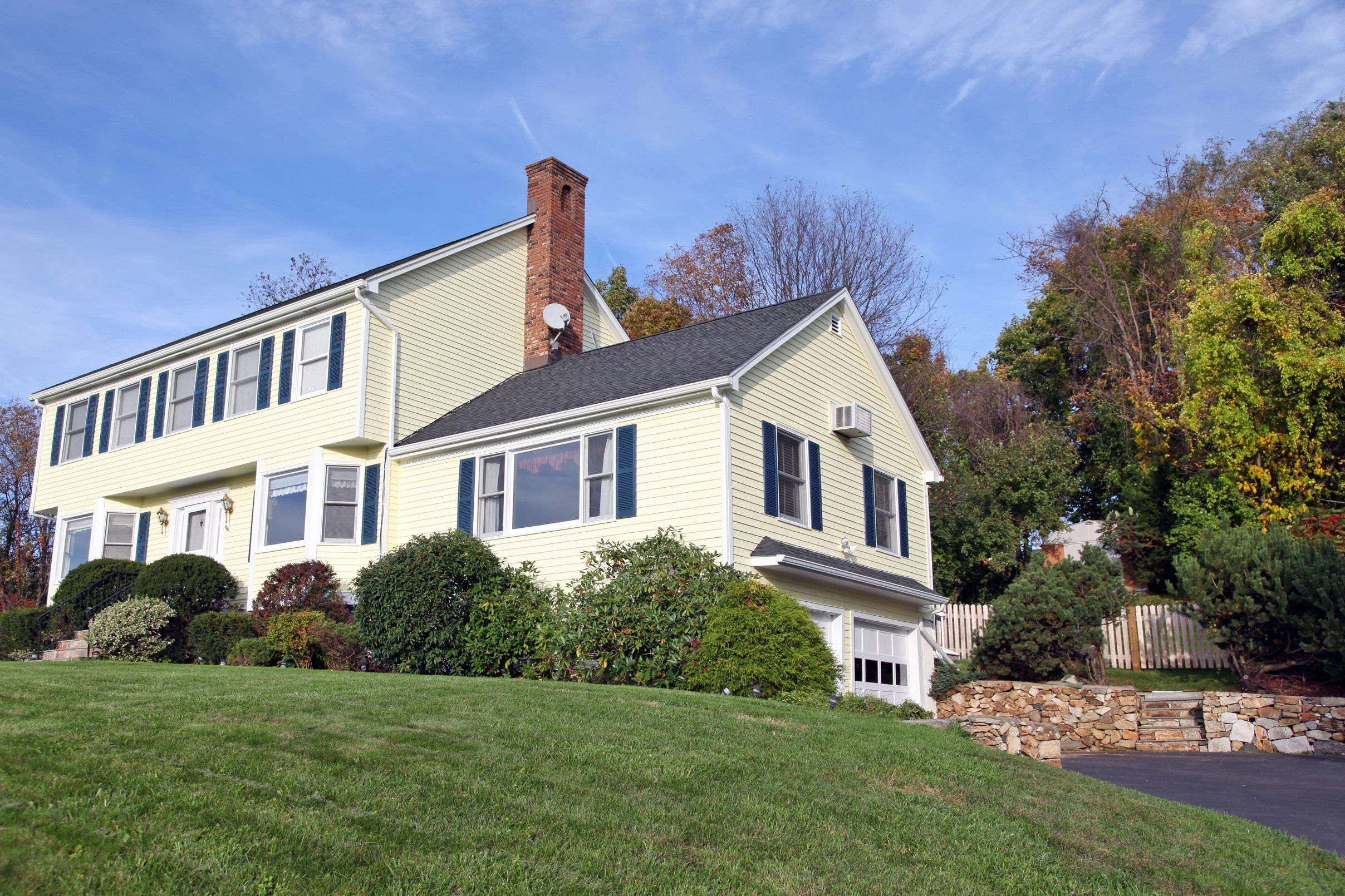 A pale yellow colonial with blue shutters, a brick chimney and a dark shingle roof on a sloping lawn.