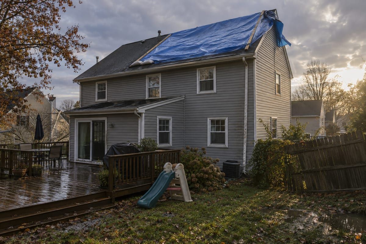 A blue tarp held down with wood strips and sandbags over the upper rear slope of a gray vinyl colonial after a storm