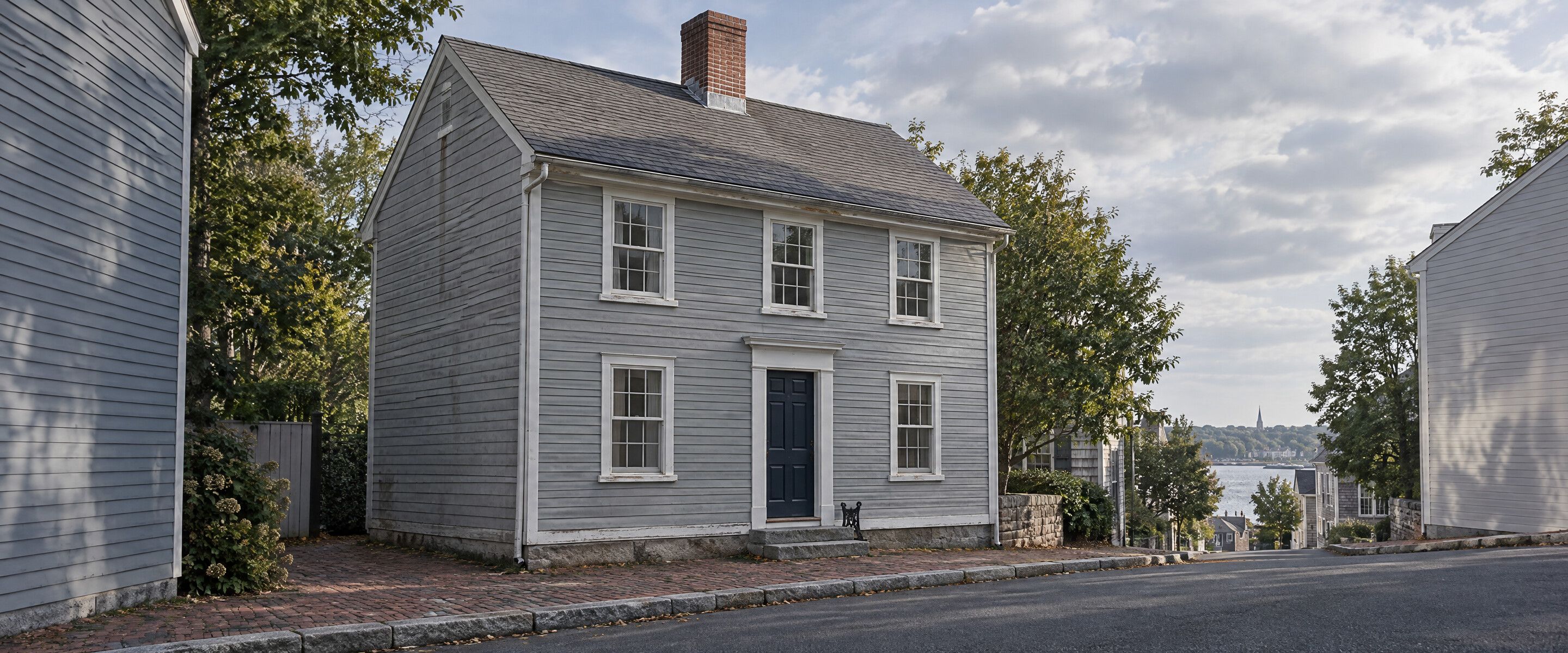 A silvered grey clapboard colonial with a charcoal shingle roof on a narrow brick-sidewalk street in an old New England town