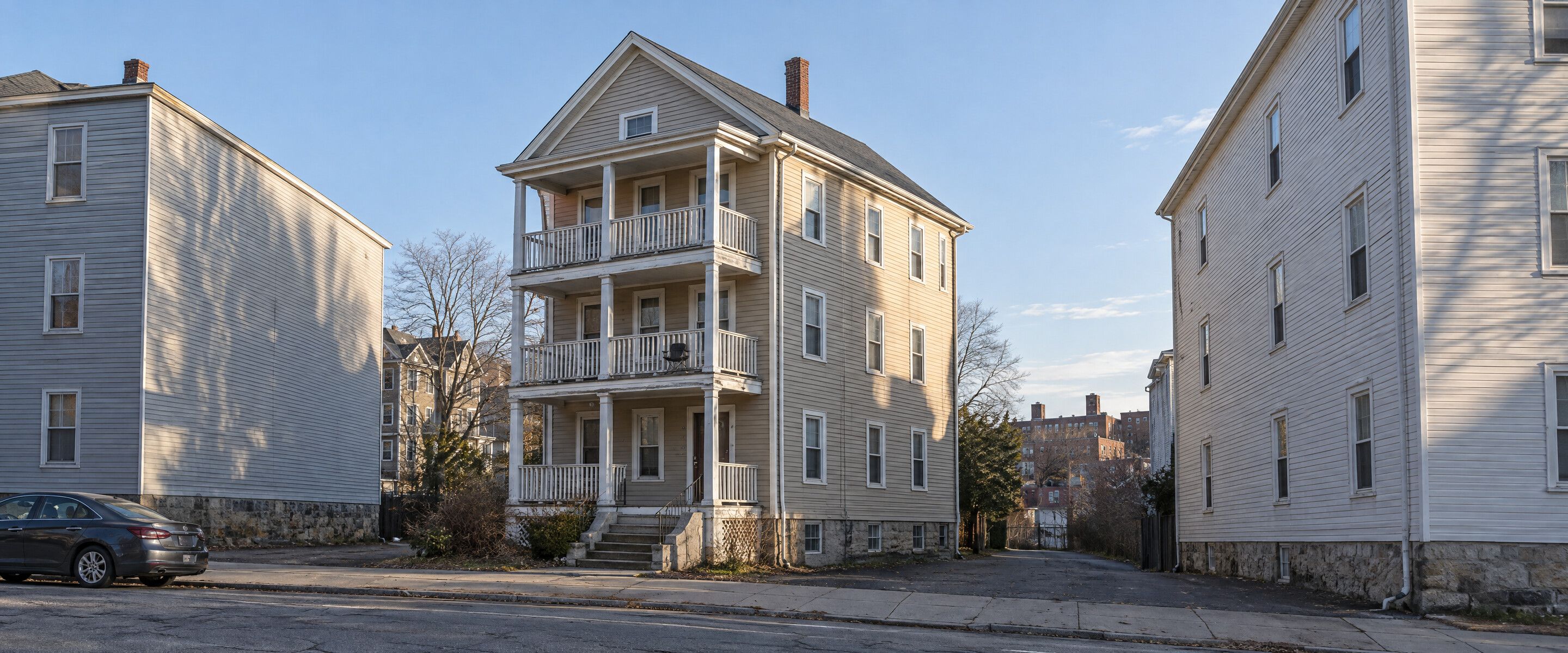 A tall wooden triple-decker with stacked open porches and putty-tan clapboard on a tight Rhode Island city street