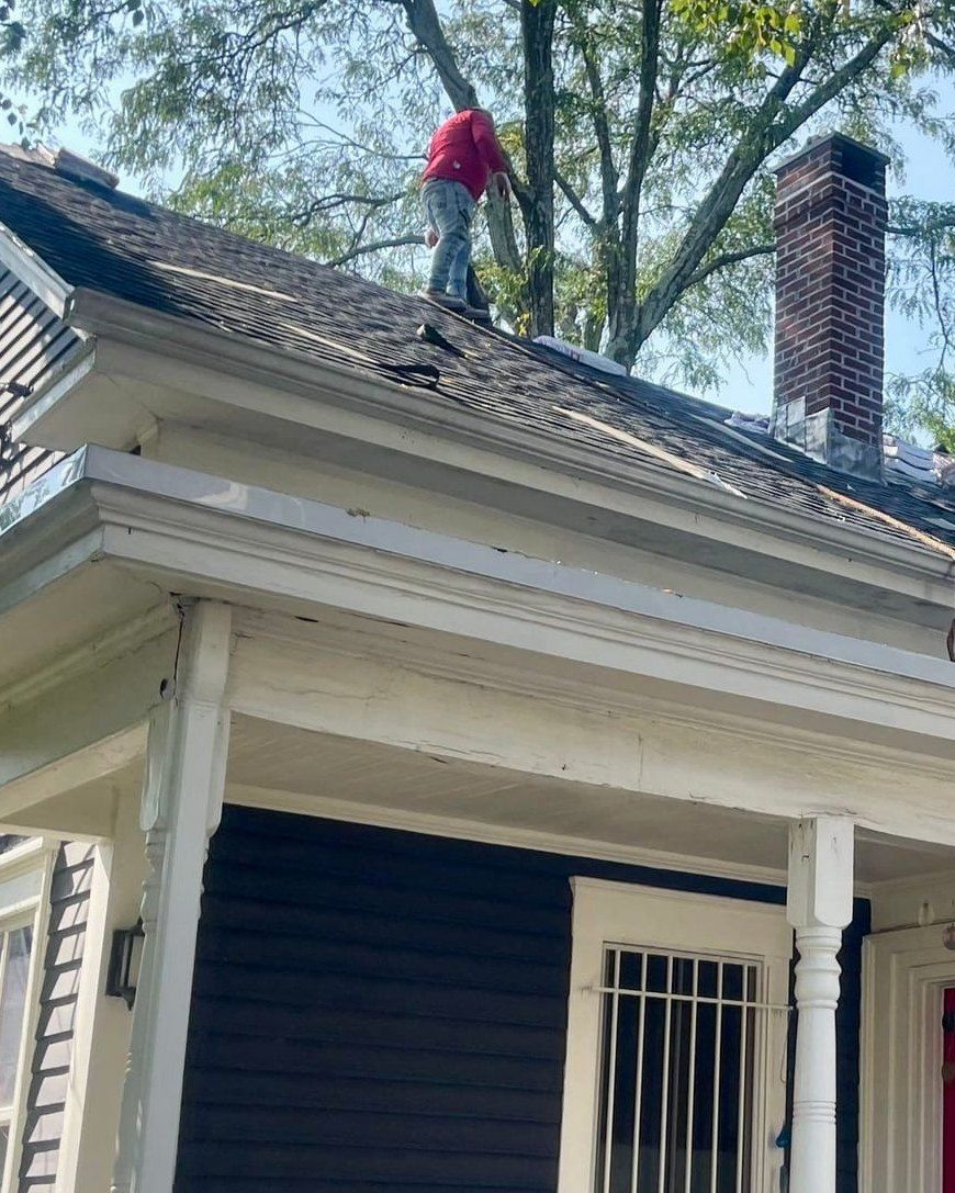Two roofers strip old shingles from the roof of a navy clapboard house with a white porch and red door.