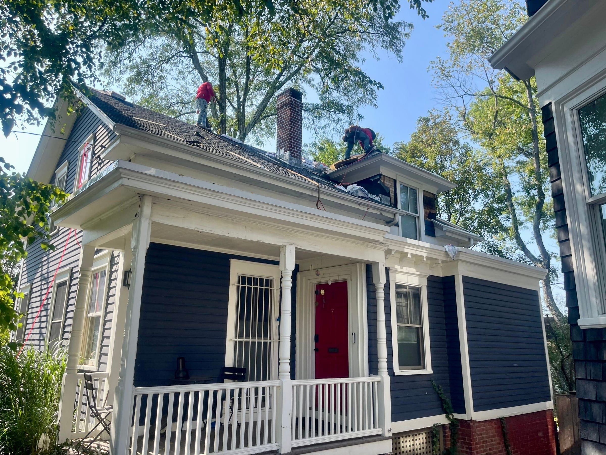 Two roofers strip old shingles from the roof of a navy clapboard house with a white porch.