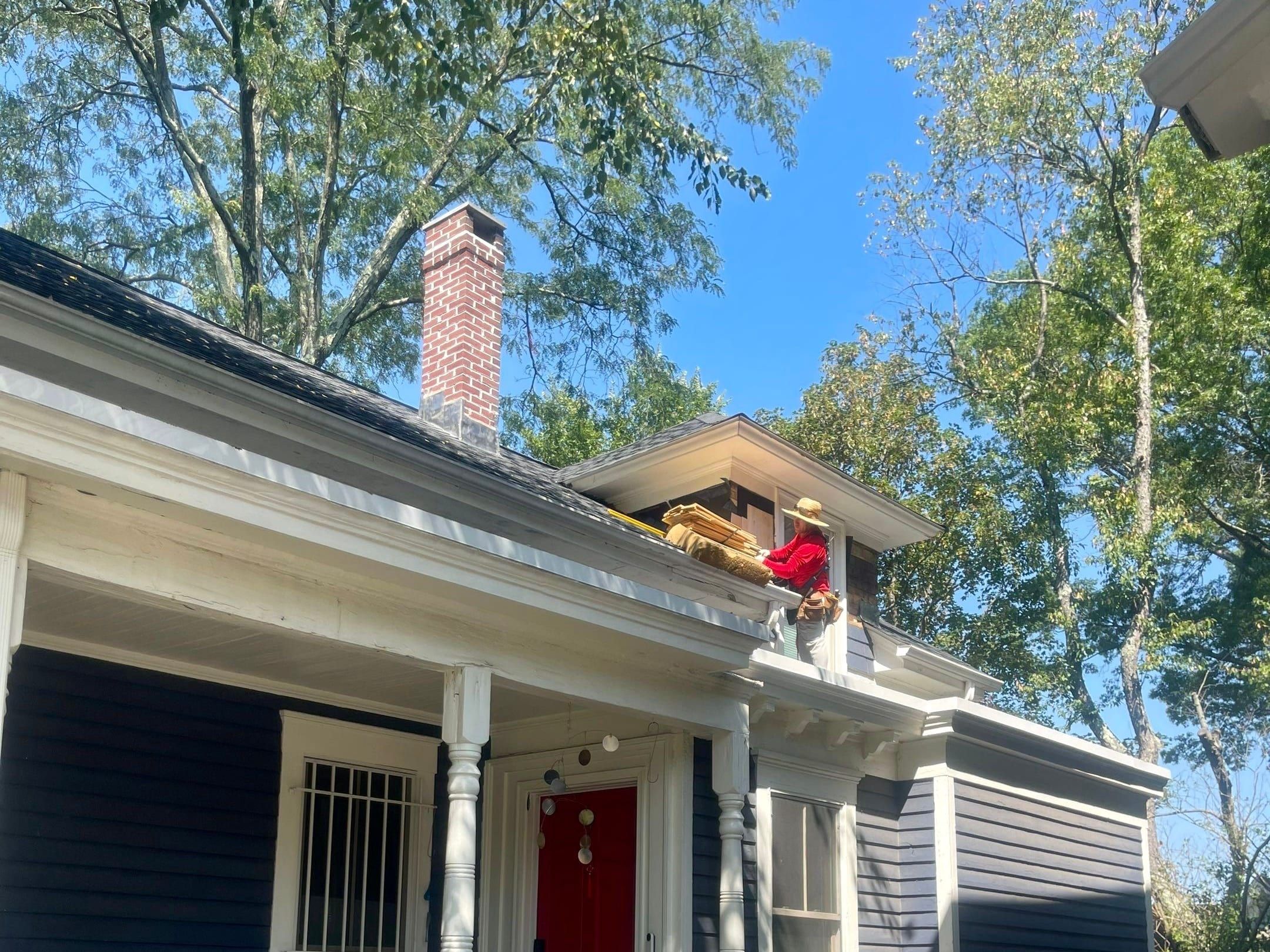 A navy clapboard house with a white porch and red door while a roofer works at the roofline above.