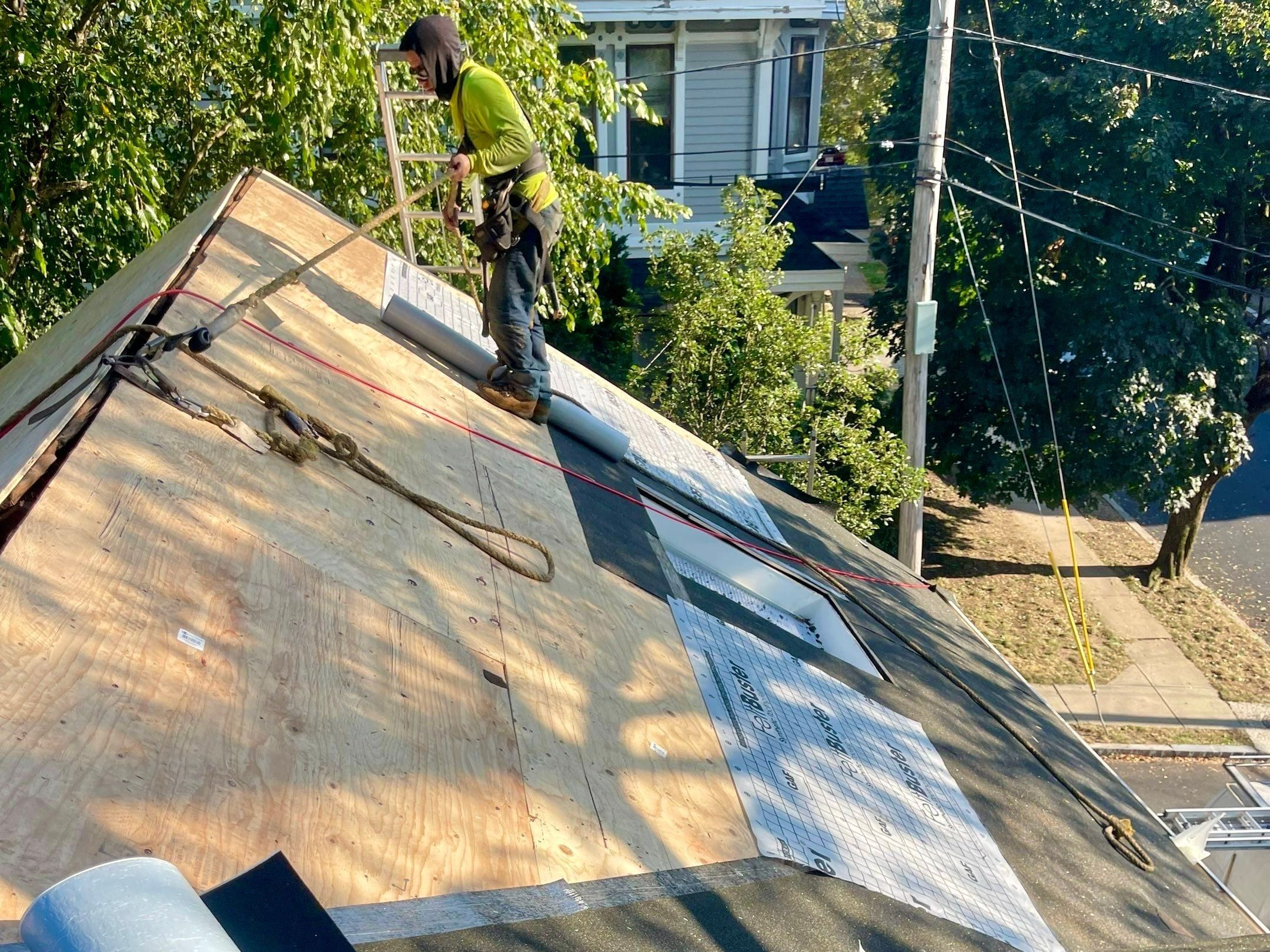 A roofer in a harness works on new plywood decking with synthetic underlayment and an ice barrier roll at the eave.