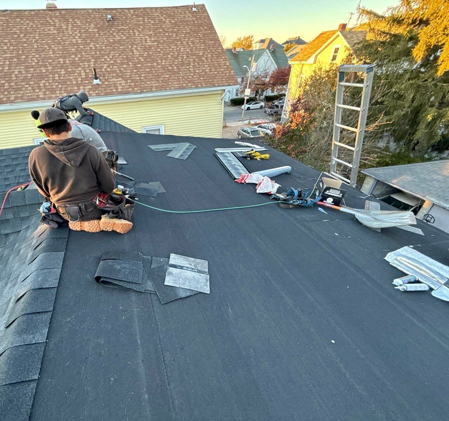 A roofer sits on a roof covered in new synthetic underlayment, with shingle scraps and tools around.