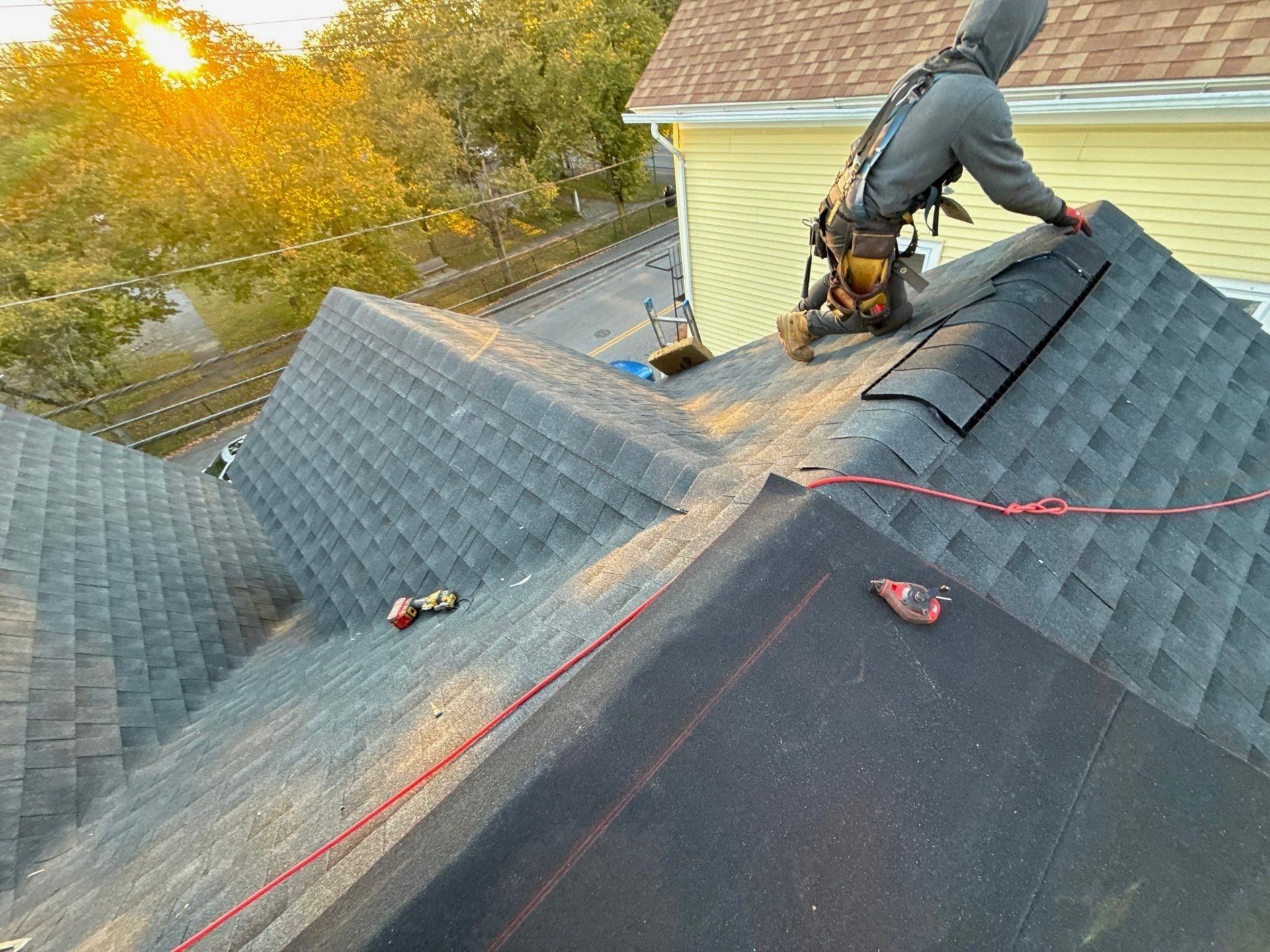 A roofer in a harness kneels at the ridge laying ridge cap on a charcoal shingle roof at sunset.
