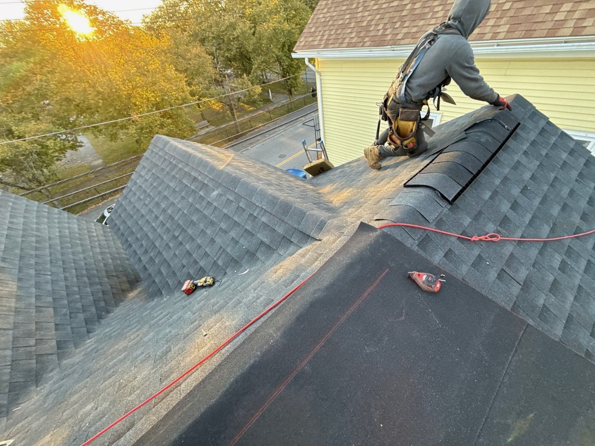 A roofer in a harness, hood up and turned away, lays ridge vent on a charcoal shingle roof at sunset.