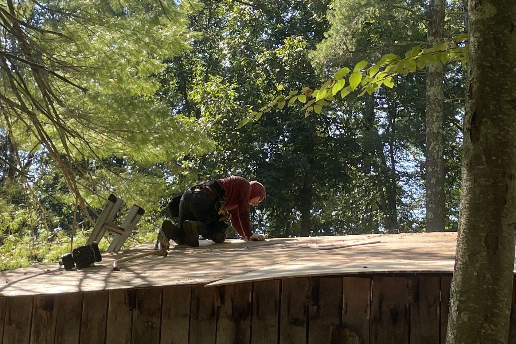 A roofer in a hooded sweatshirt kneels on new plywood sheathing on an outbuilding roof under tall trees.