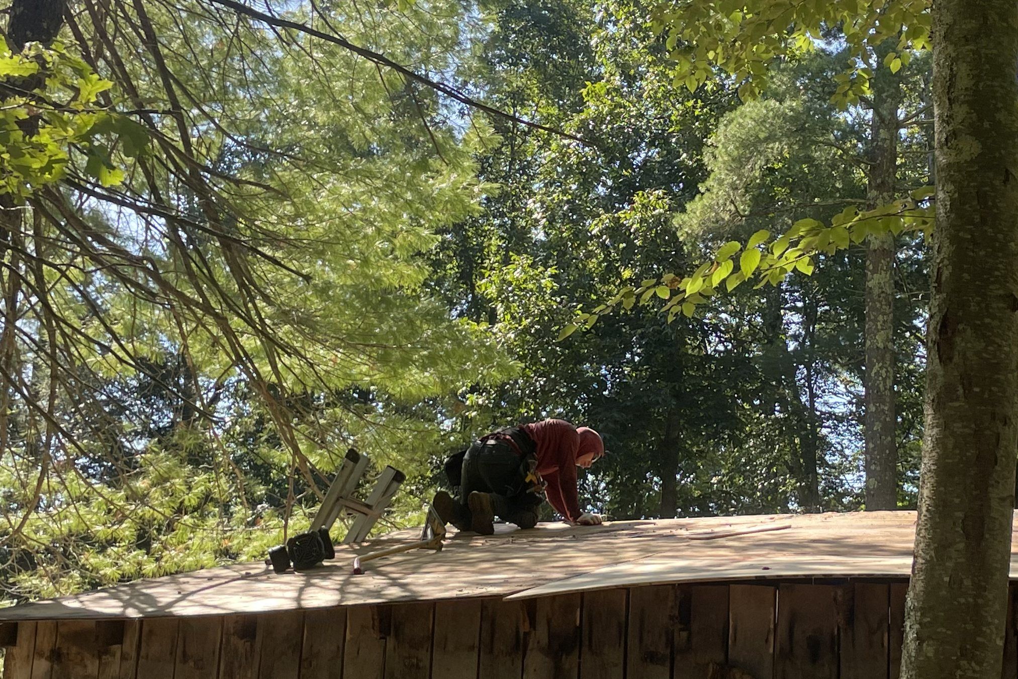 A roofer in a hooded sweatshirt kneels on new plywood sheathing on an outbuilding roof under tall trees.