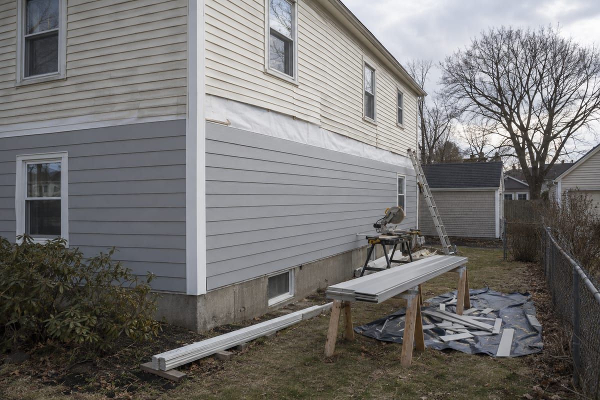 A colonial side wall mid re-siding, new pale grey lap siding below old cream vinyl, with a saw and new boards in the yard.