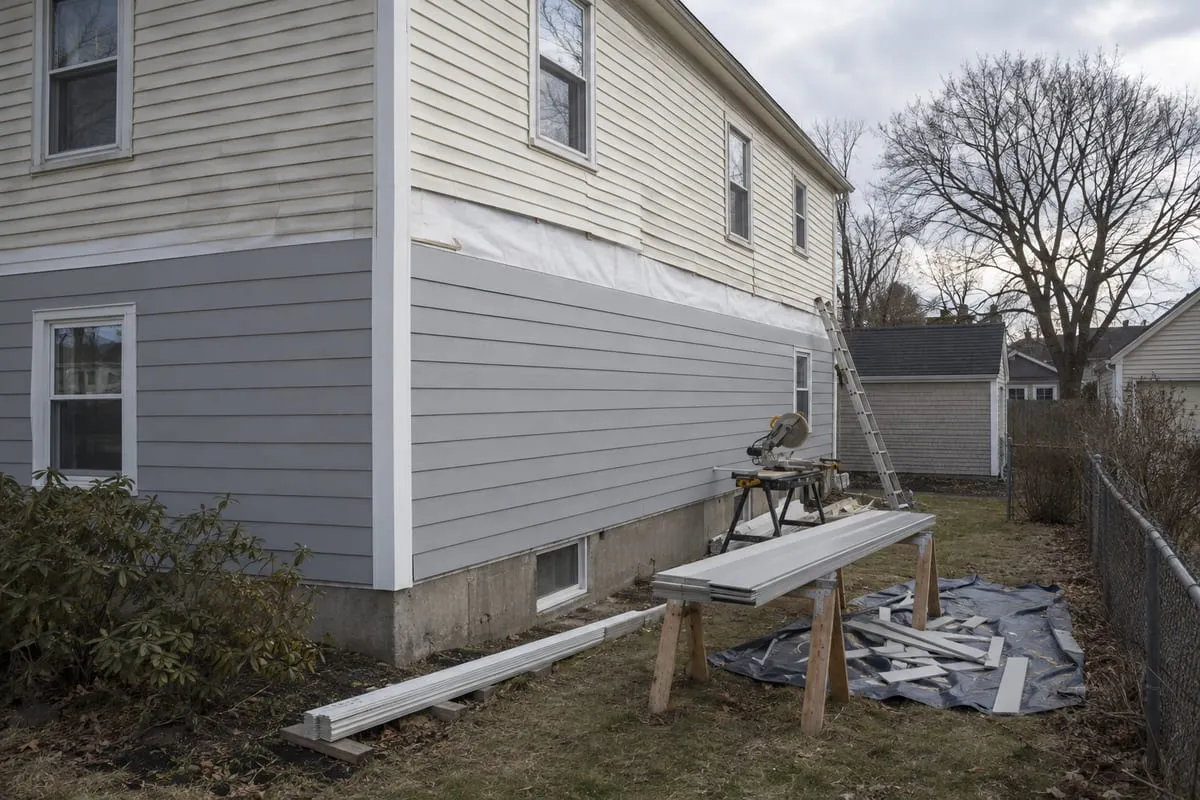 A colonial side wall mid siding replacement, new pale grey lap siding below old cream vinyl, with a saw and new boards in the yard