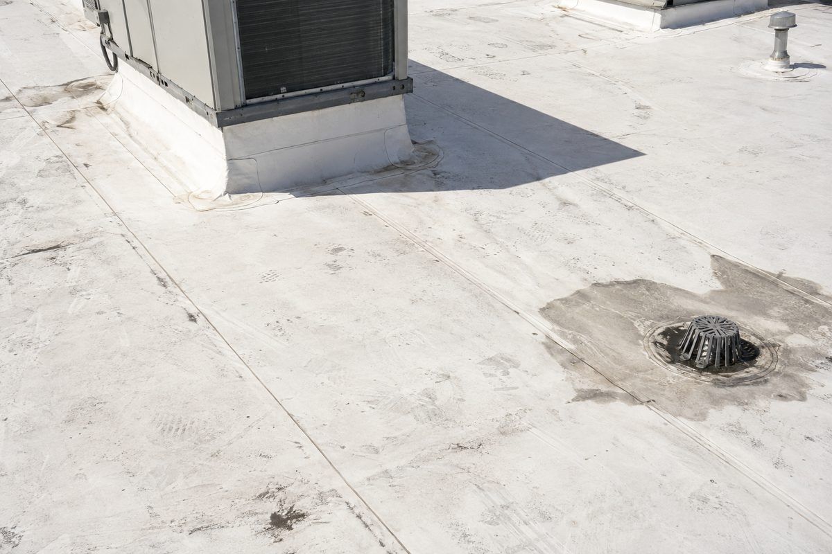 Close three-quarter view of a scuffed white single-ply roof with a rooftop unit curb, a vent stack and a drain in a dirty pond ring